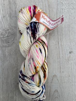 Ein Strang handgefärbte Wolle, Herzfasern No. 13 „Sprenkelparty“. Die Basis ist naturweiß, darüber verteilt bunte Farbsprenkel in Pink, Gelb, Blau, Violett und Schwarz. Der Strang liegt gedreht auf hellem Holz, das Herzfasern-Etikett ist sichtbar angebrac